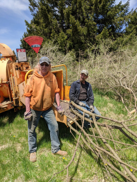 Two men in work clothes pose near a wood chipping machine and scrubby trees.