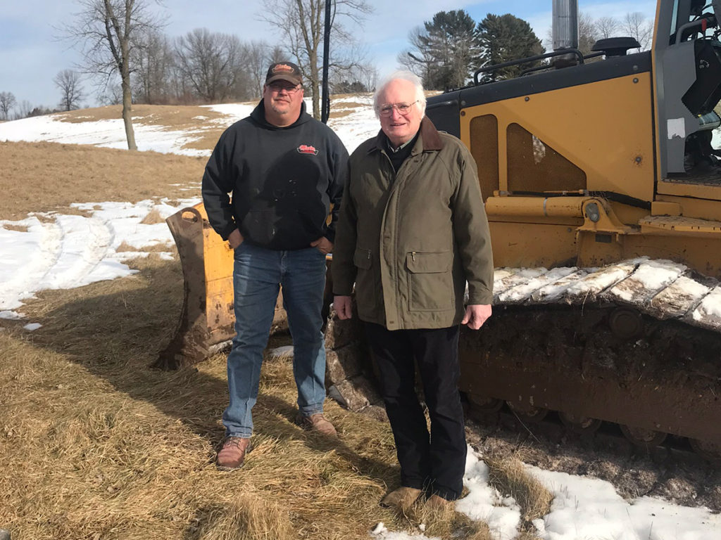 a man in a sweatshirt and cap and a man in a coat stand in front of a bulldozer on a winter day