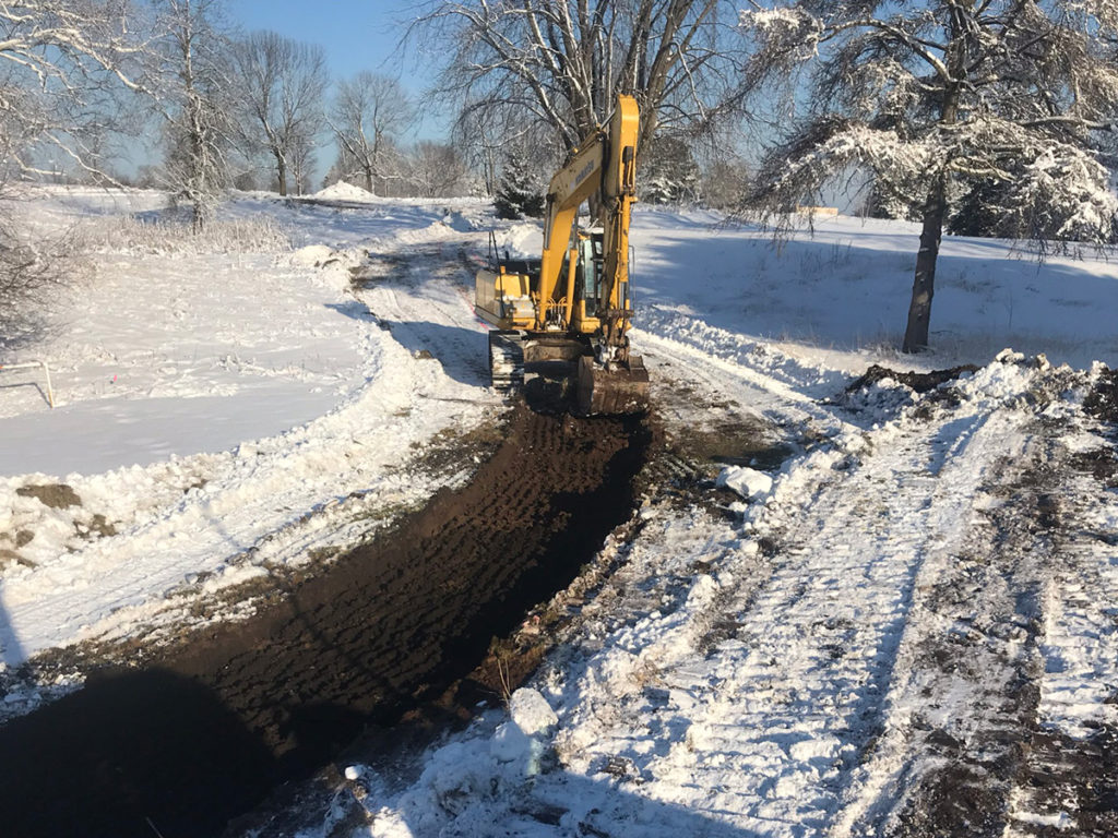 clear dirt path in snowy field with excavation vehicles in background and a row of homes