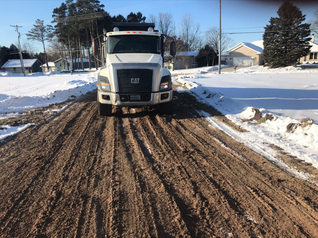 front view of a white dump truck drives on a cleared dirt path through snow covered field