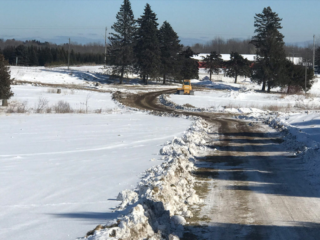 a snow covered field with hills and trees in the background and a cleared drive in the foreground