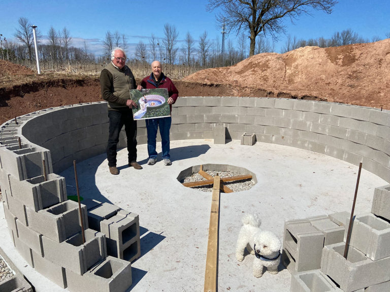 Two men and a small white dog stand outdoors on a sunken concrete circle surrounded by block retaining walls. The men hold a poster of what the finished structure will look like.