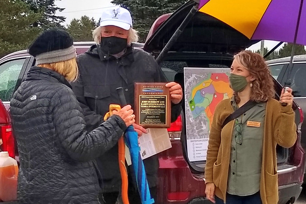 Two women stand on either side of a man holding a plaque. It is raining.