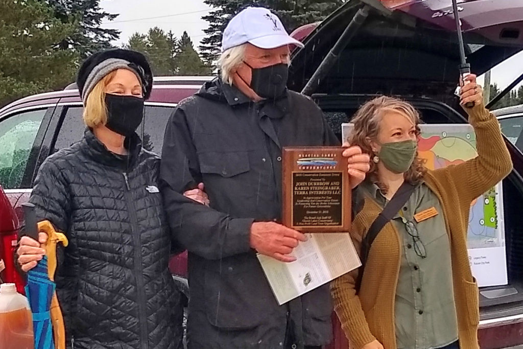 Two women stand on either side of a man holding a plaque. It is raining.