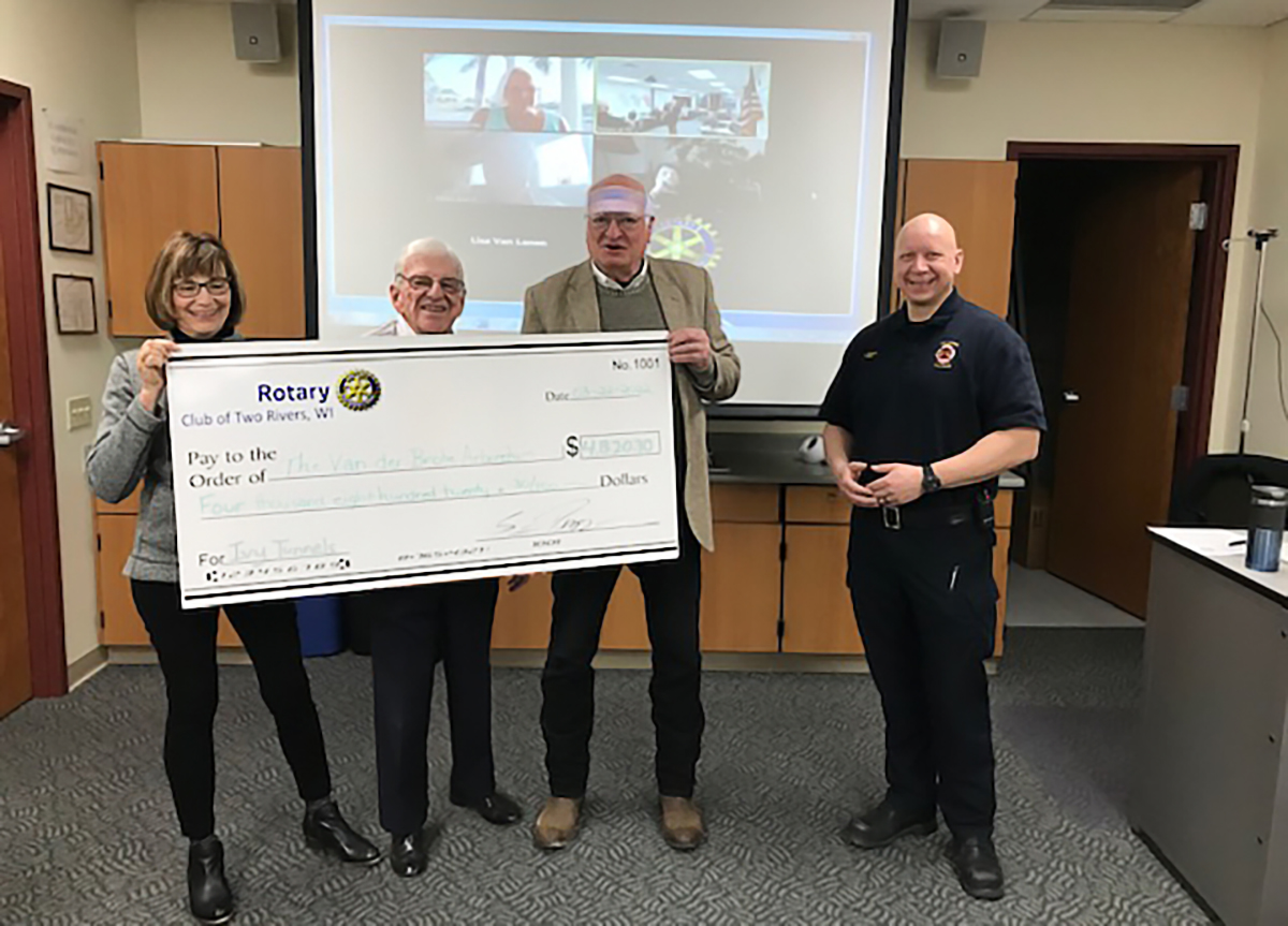 4 people in a meeting room. One woman and two men stand behind and hold an oversized check for $4820.30. A fourth man is dressed in a police officer uniform.