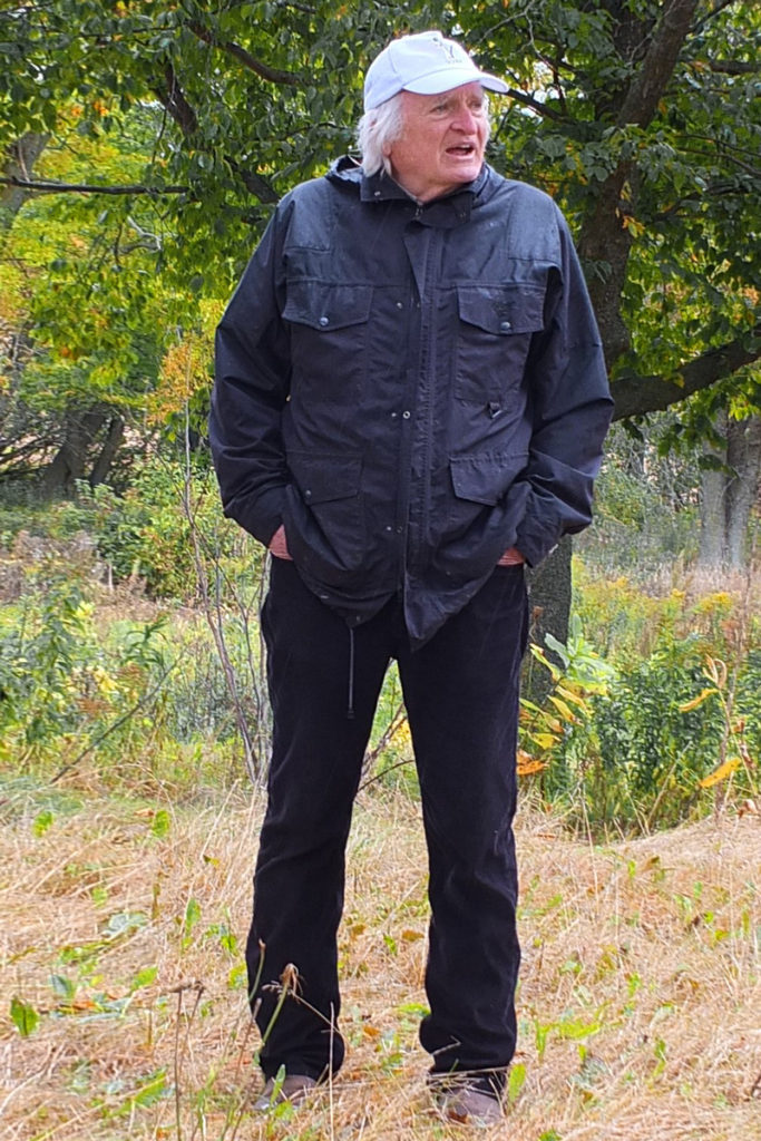 A man with white hair and wearing a jacket and cap speaks outdoors.