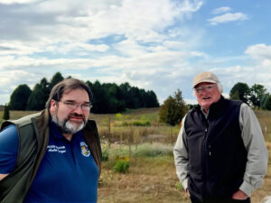 two men in a meadow on a bright day. One wears a cap and one wears a state senator t-shirt.