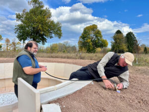 two men in an outdoor meeting circle made of stone fiddling with a gas line