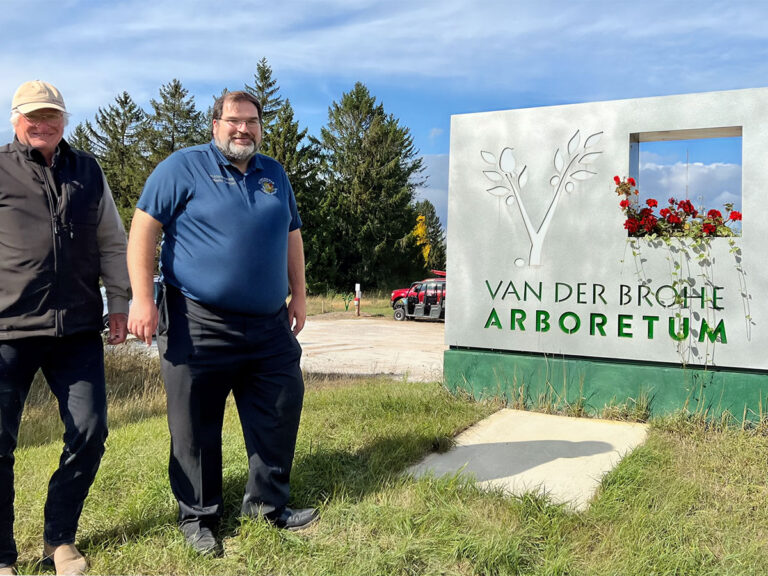 two men in front of a large lighted metal sign reading vanderbrohe arboretum