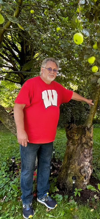 A mature man in a red t-shirt with a white W stands in front of an apple tree with many large yellow apple.