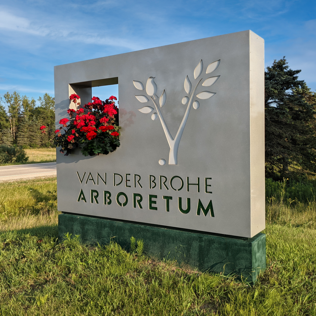 a large gray and green metal entrance sign with a geranium planter built in and the VDBA logo cut out of the metal