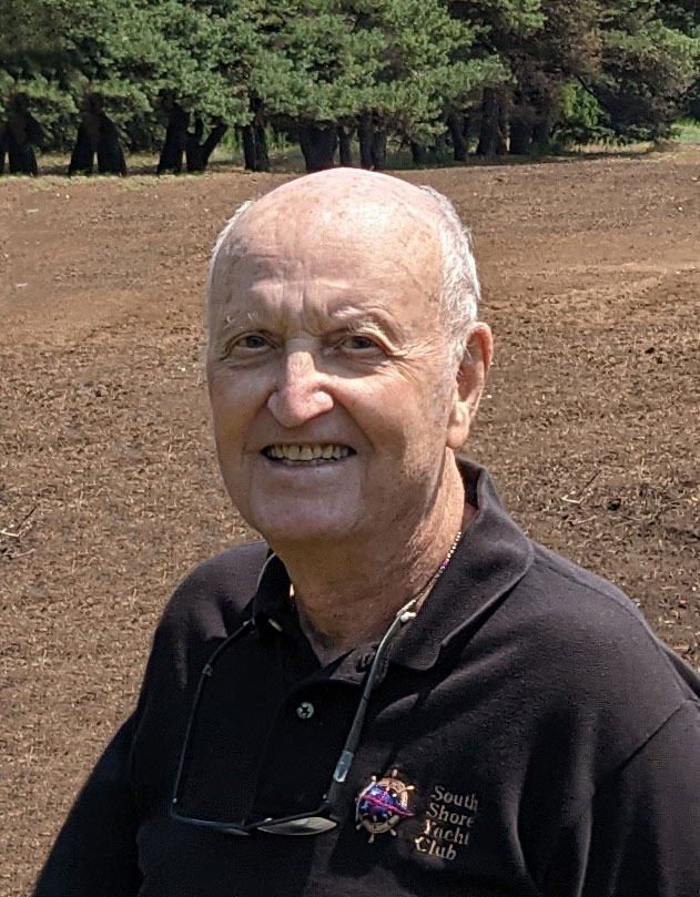 an older man with white hair and a black polo shirt stands in front of a bare meadow with trees in the background.