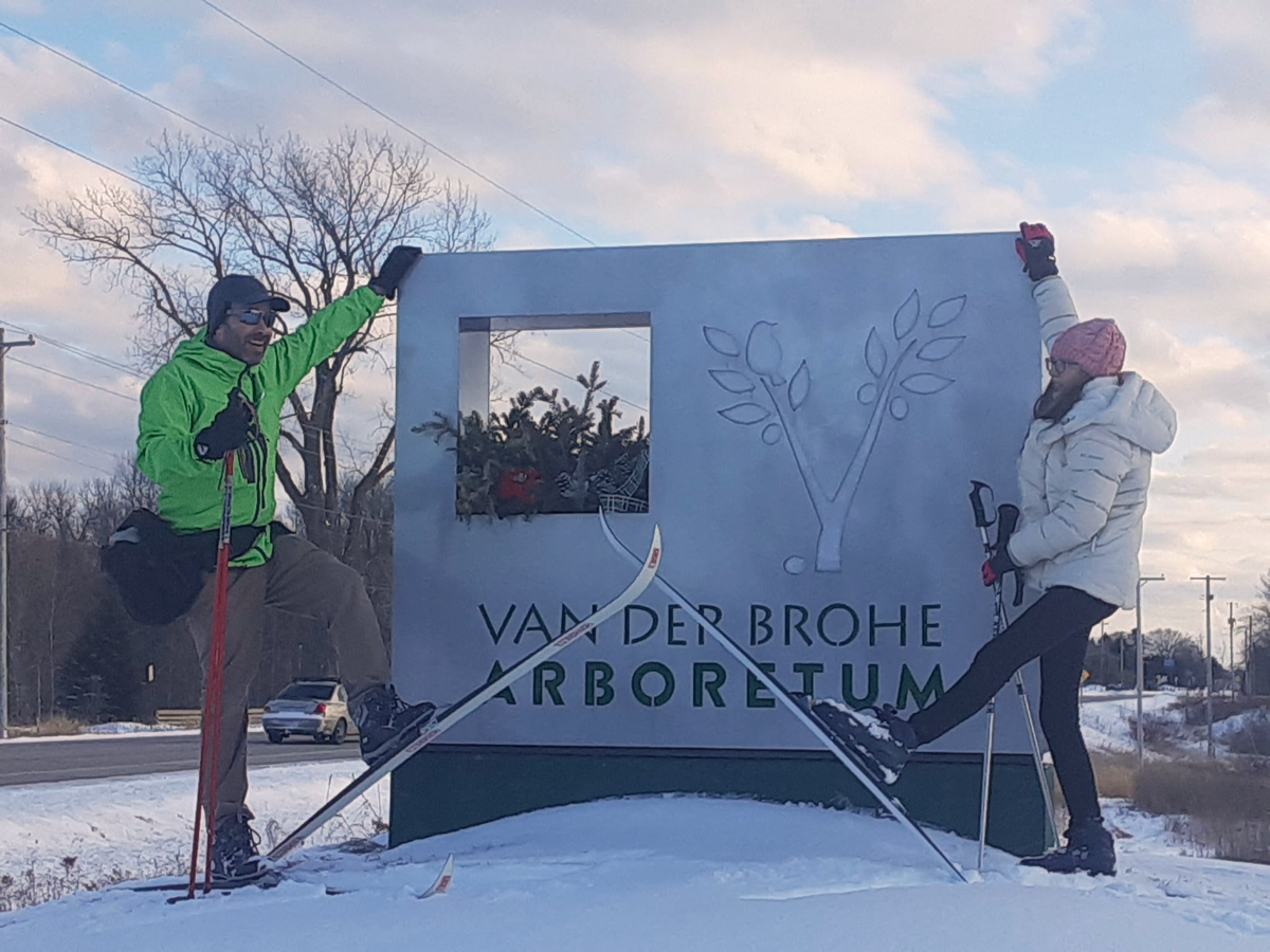 A man and a woman pose with their cross-country skiis crossed in front of the Arboretum entrance sign.