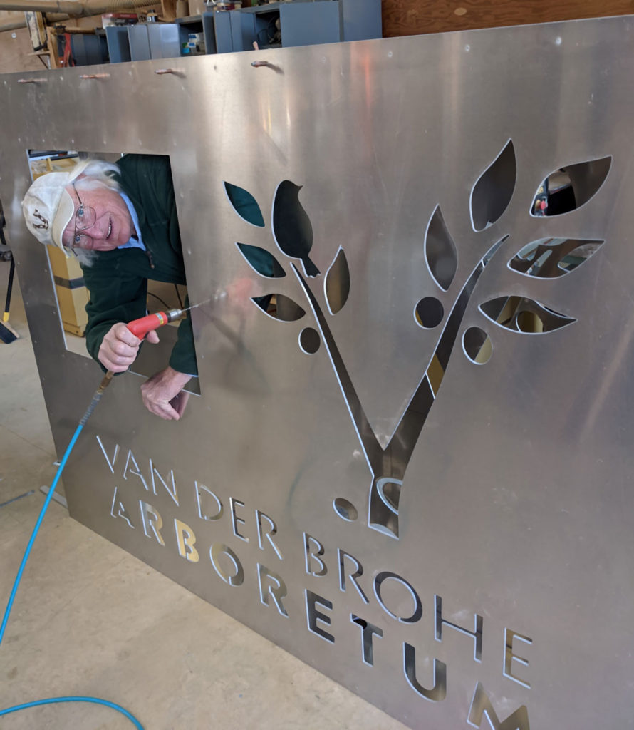 A man with white hair and a cap drills on a large aluminum sign while peeking through a cutout in the sign. The sign also has a tree and bird cutout, as well as the worlds VAN DER BROWH ARBORETUM.
