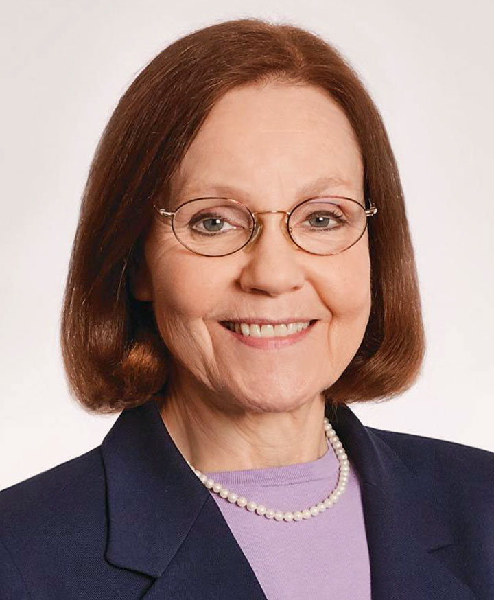 An older caucasian female with auburn hair in a bob, glasses, a suit with lavender blouse and pearl necklace.