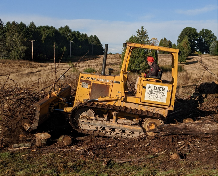 a yellow bulldozer with F. Dier printed on side driven over dirt by a man. There are trees in the background.