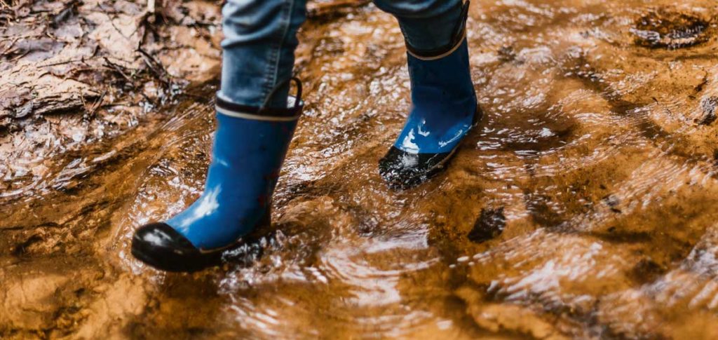 a person with blue rubber boots is shown from the knee down, walking through water and mud.